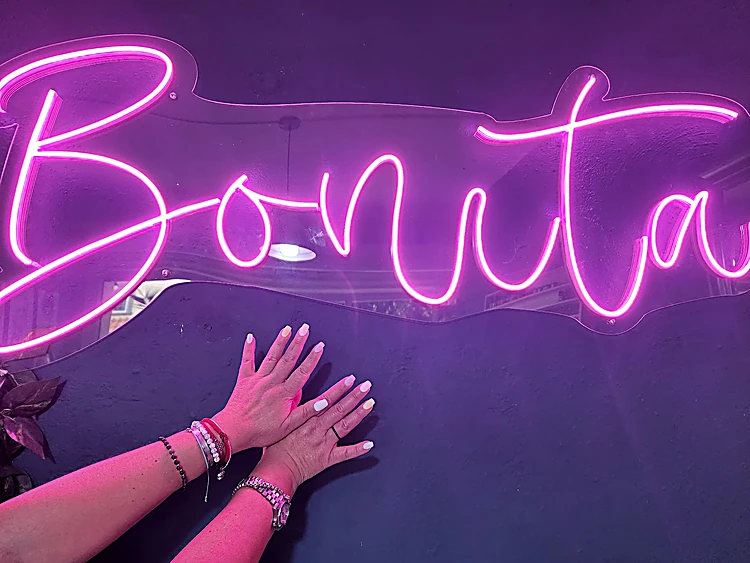 Manos de una clienta con uñas pastel recargadas en el muro, bajo el neón Bonita