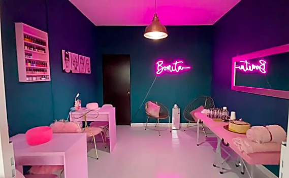 Interior del salón: muros azul petróleo, mesas de manicura rosas, lámpara colgante y dos letreros de neón Bonita