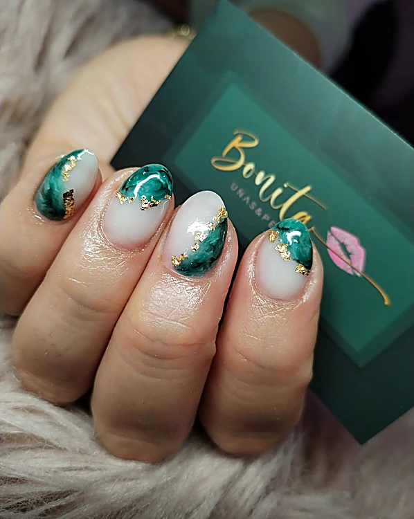 Uñas cortas en verde esmeralda marmoleado con hoja de oro, sobre peluche rosa, junto a la tarjeta teal de Bonita Uñas y Pestañas